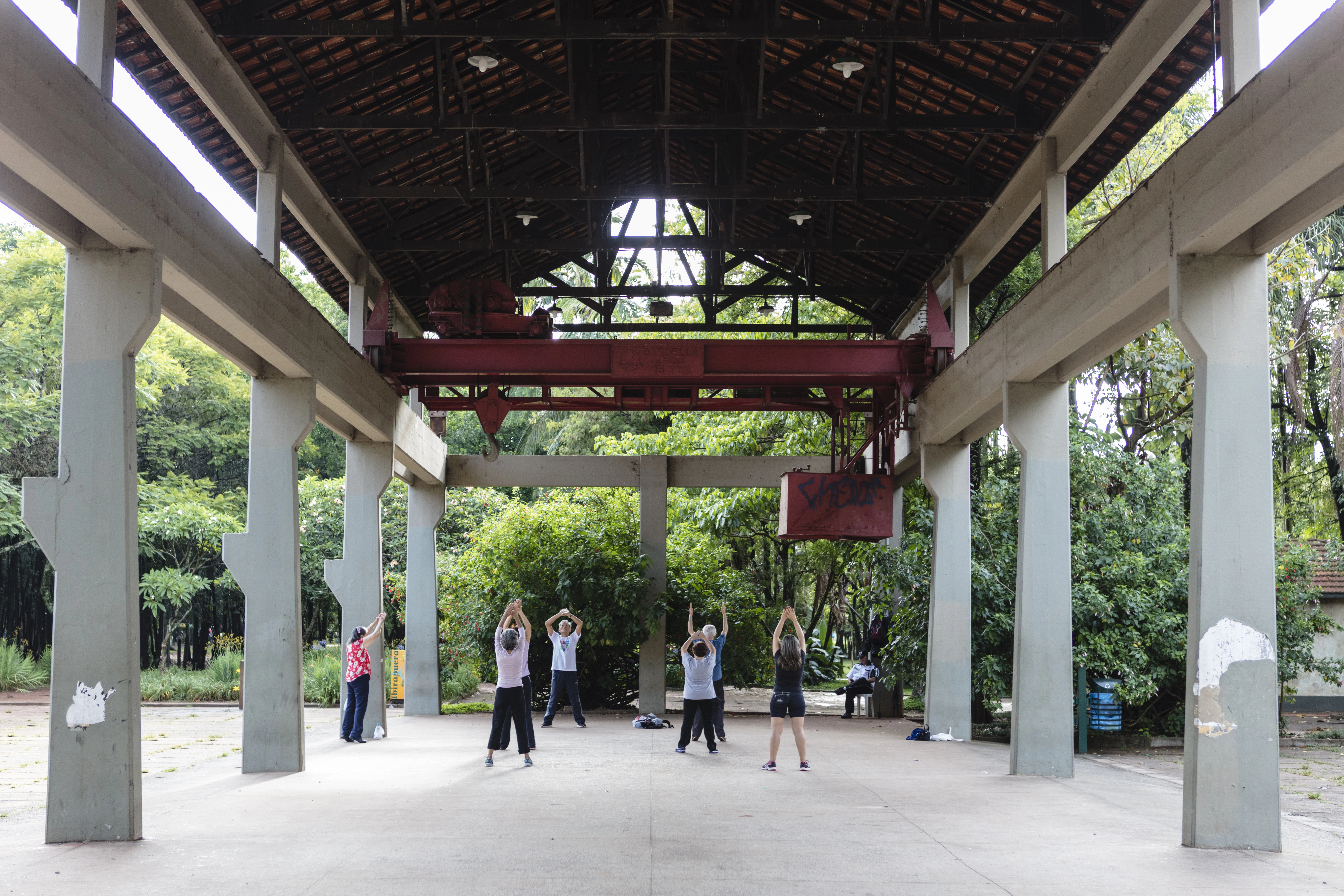 Foto de uma estrutura externa, um corredor de concreto e madeira bastante aberto. Nele, algumas pessoas circulam e atrás há uma parede de árvores. 